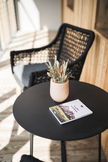Wellnesshotel in Südtirol: Auf in den Traumurlaub! Schwarzer runder Tisch mit Trockenblumen und Broschüre, schwarzer Stuhl im Hintergrund