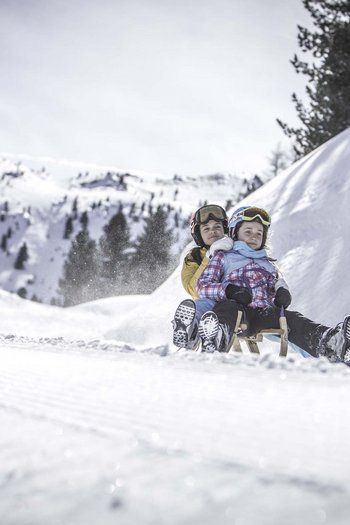 Il nostro hotel sulle piste a San Vigilio di Marebbe Due bambini che fanno slittino insieme su una collina innevata con montagne sullo sfondo