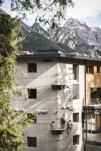Wellnesshotel in Südtirol: Auf in den Traumurlaub! Menschen klettern an einer Kletterwand an einem modernen Gebäude mit Bergen im Hintergrund