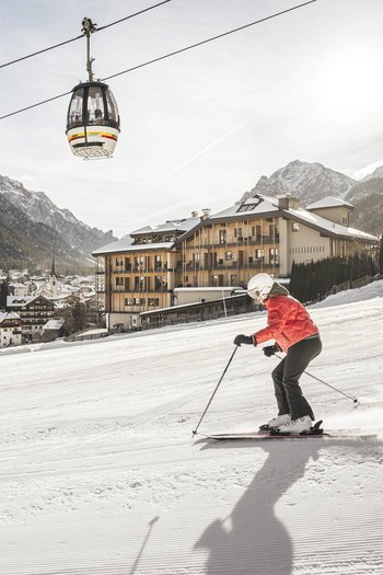 Wellnesshotel in Südtirol: Auf in den Traumurlaub! Skifahrer mit roter Jacke auf Piste vor Bergdorf und Gondeln im Schnee