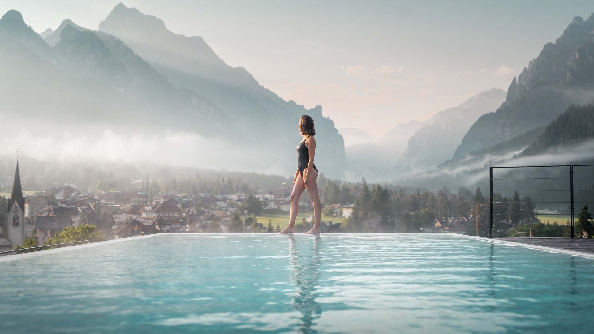 Angebote: Wellness und Wandern | Südtirol Frau im Badeanzug steht am Rand eines Pools mit Bergblick und Dorf