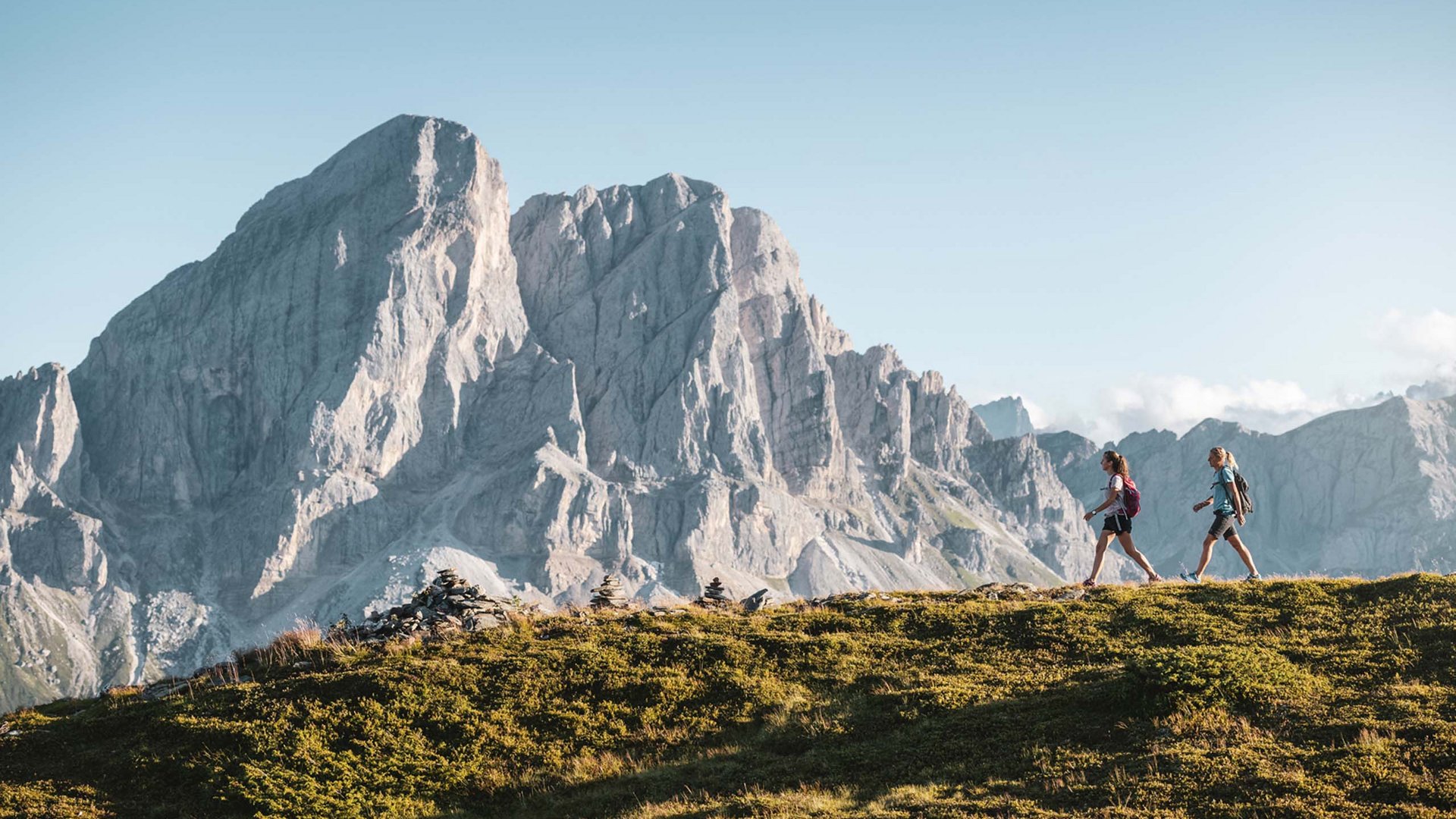 Angebote: Wellness und Wandern | Südtirol Zwei Wanderer wandern auf einem grasbewachsenen Hügel mit Berg im Hintergrund